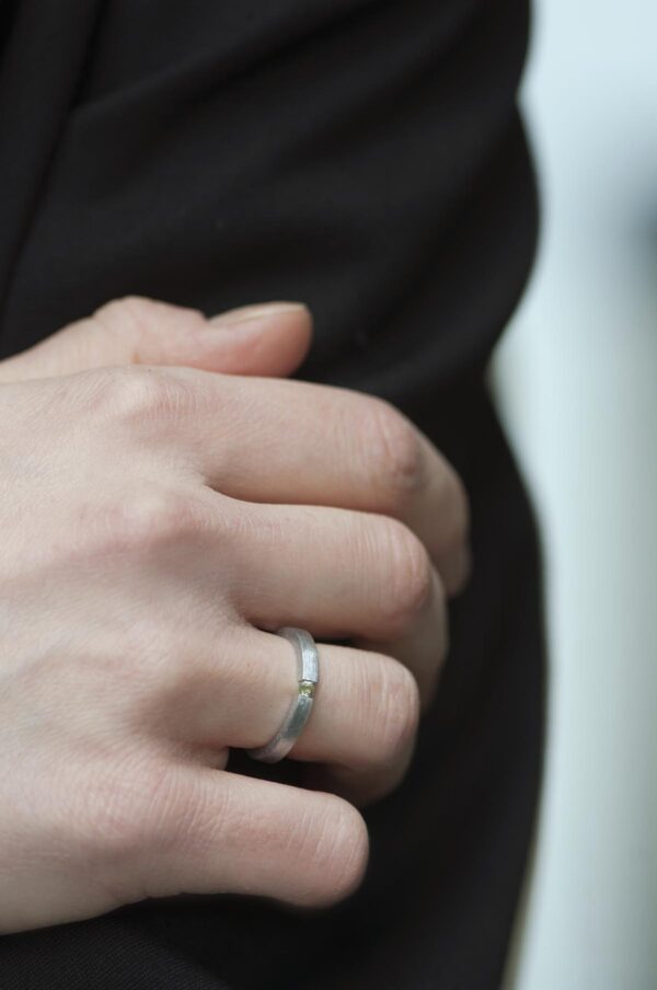 Minimalistyczny nowoczesny pierścionek ze srebra z jasnozielonym perydotem założony na serdeczny palec. Prosta obrączka o prostokątnym przekroju ma szerokość i wysokość kamienia. Końce obrączki ściskają z obu stron kamień. Pierścionek pokazany od boku. Wyraźnie widoczny typ oprawy tension ring. Wykończenie satynowe.