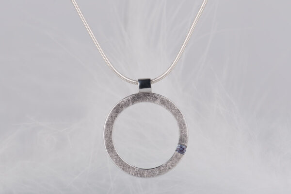 Okrągła srebrna zawieszka z aleksandrytem, zawieszona na łańcuszku typu linka. Kamień jest ustawiony asymetrycznie na godzinie czwartej.