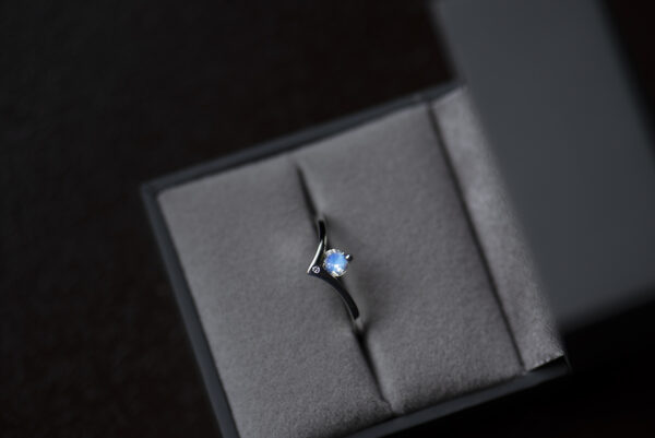 Pierścionek z białego złota 585 z niebieskim kamieniem księżycowym umieszczony w szarym pudełku na biżuterię. Szyna wygięta w kształcie litery v. Okrągły kamień oprawiony w miejscu wcięcia. Poniżej w szynie na płasko oprawiony mały diament.