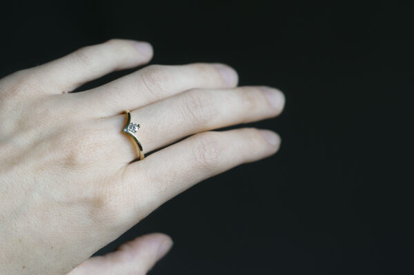 Pierścionek z żółtego złota z syntetycznym białym moissanitem 0,3 ct. Szyna ugięta w kształcie litery V. Kamień oprawiony we wcięciu. Poniżej oprawiony w szynie na płasko naturalny mały diament. Pierścionek założony na środkowy palec.