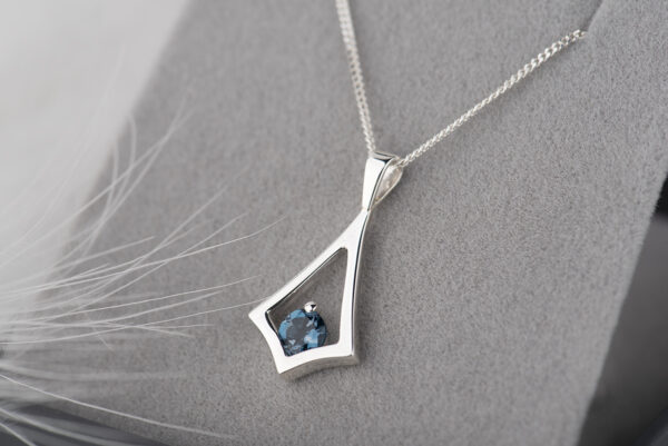 Srebrna zawieszka w kształcie latawca z okrągłym topazem London blue oprawionym w dolnej części. Zawieszka wisi na łańcuszku typu pancerka, którego fragment widać na zdjęciu. Zawieszka umieszczona w szarym pudełku na biżuterię.