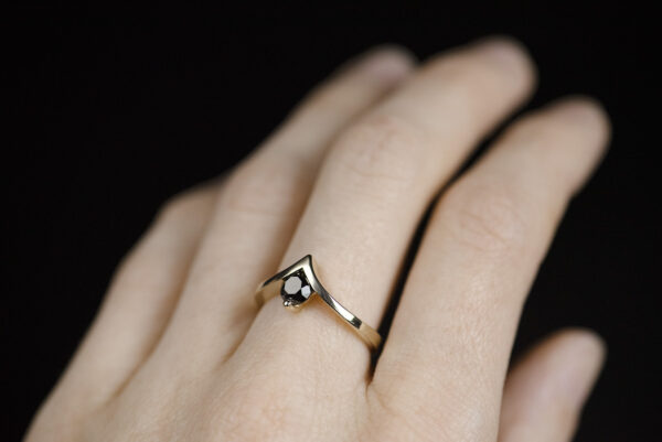 Pierścionek z żółtego złota z czarnym diamentem 0,5 ct założony na palec. Szyna pierścionka jest wygięta w kształcie litery v. W miejscu wcięcia jest oprawiony czarny kamień.