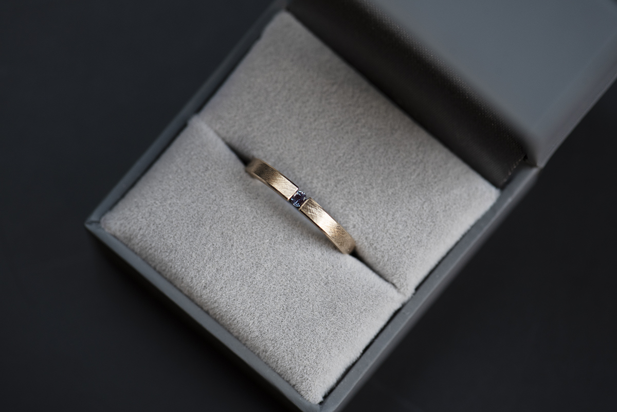 Minimalistyczny pierścionek typu tension ring z żółtego złota próby 585 z syntetycznym zmieniającym kolor aleksandrytem. Końce prostej szyny o przekroju prostokątnym ściskają kamień z obu stron. Pierścionek przypomina prostą obrączkę. Może pełnić funkcję pierścionka zaręczynowego dla kobiety lub mężczyzny lub obrączki. Pierścionek włożony do szarego pudełka na biżuterię pokazany z góry.