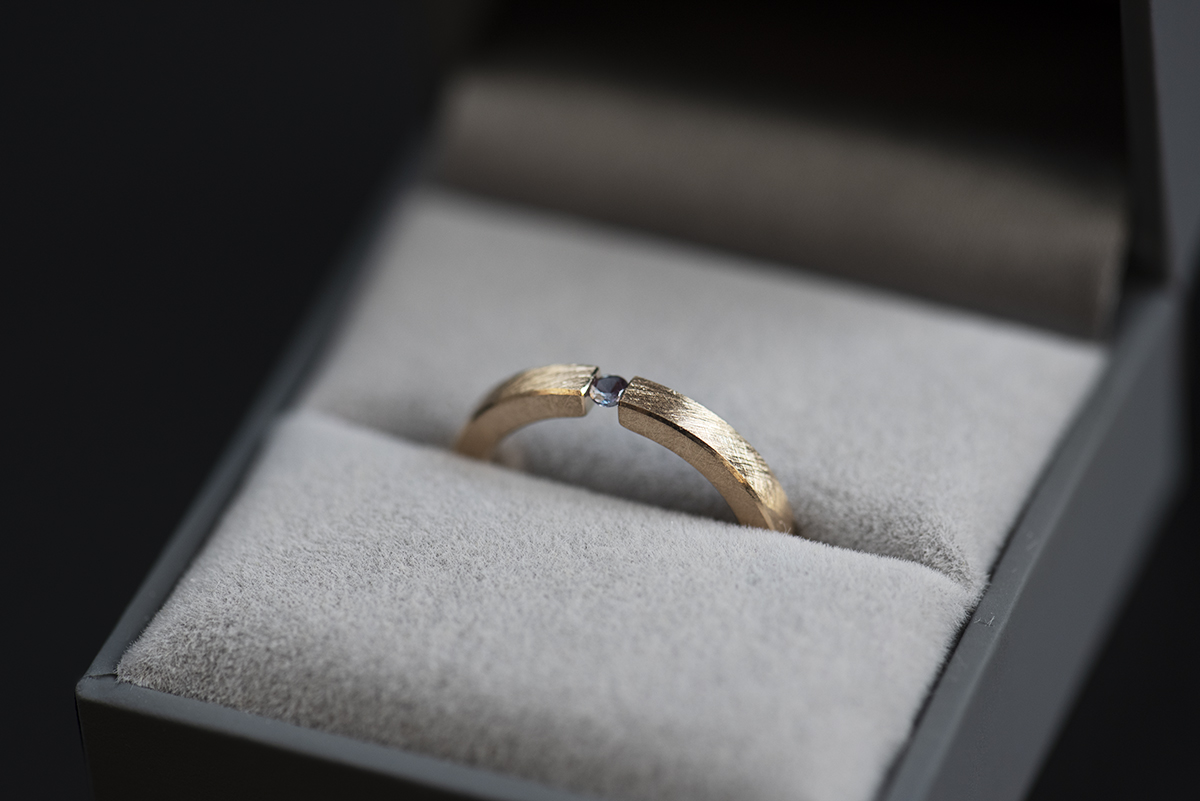 Minimalistyczny pierścionek typu tension ring z żółtego złota próby 585 z syntetycznym zmieniającym kolor aleksandrytem. Końce prostej szyny o przekroju prostokątnym ściskają kamień z obu stron. Pierścionek przypomina prostą obrączkę. Może pełnić funkcję pierścionka zaręczynowego dla kobiety lub mężczyzny lub obrączki. Pierścionek włożony do szarego pudełka na biżuterię pokazany z góry.