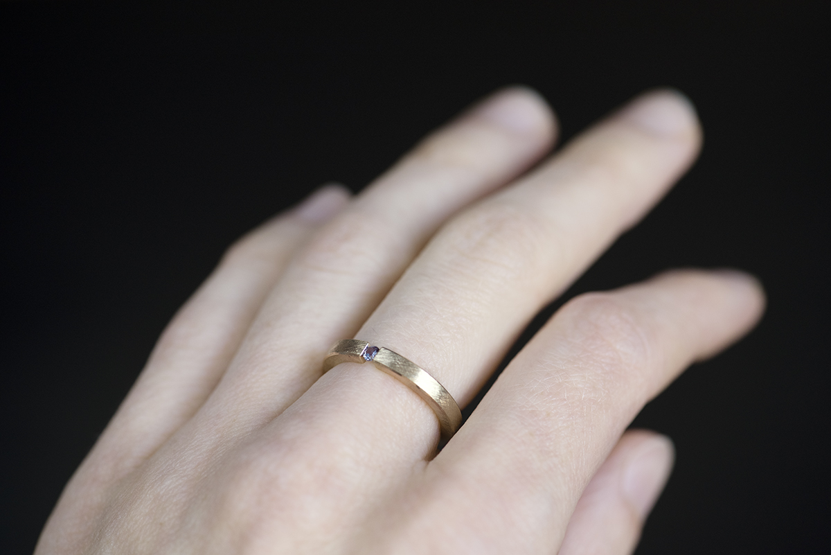 Minimalistyczny pierścionek typu tension ring z żółtego złota próby 585 z syntetycznym zmieniającym kolor aleksandrytem. Końce prostej szyny o przekroju prostokątnym ściskają kamień z obu stron. Pierścionek przypomina prostą obrączkę. Może pełnić funkcję pierścionka zaręczynowego dla kobiety lub mężczyzny lub obrączki. Pierścionek założony na palec środkowy.