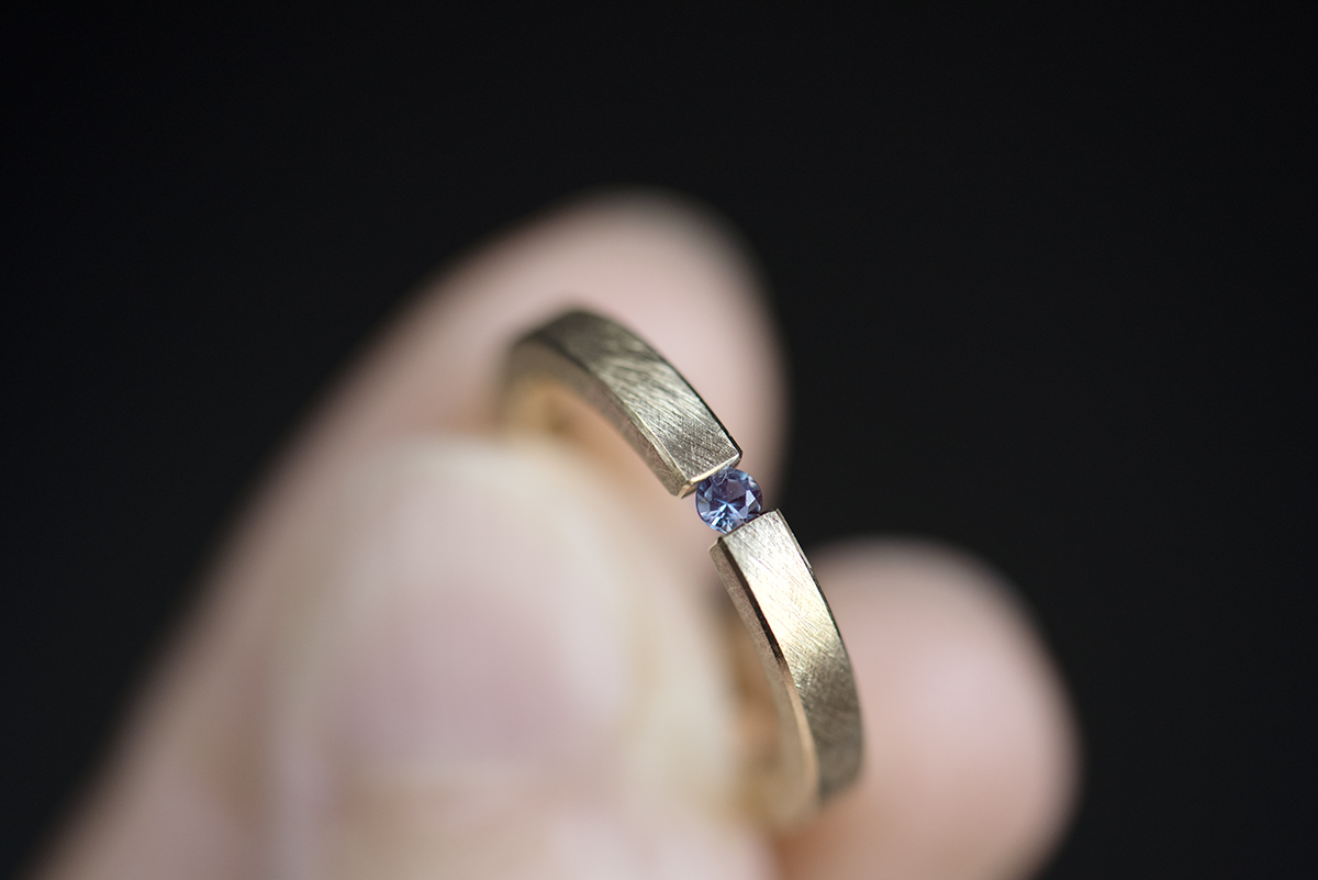 Minimalistyczny pierścionek typu tension ring z żółtego złota próby 585 z syntetycznym zmieniającym kolor aleksandrytem. Końce prostej szyny o przekroju prostokątnym ściskają kamień z obu stron. Pierścionek przypomina prostą obrączkę. Może pełnić funkcję pierścionka zaręczynowego dla kobiety lub mężczyzny lub obrączki. Pierścionek trzymany w palcach.
