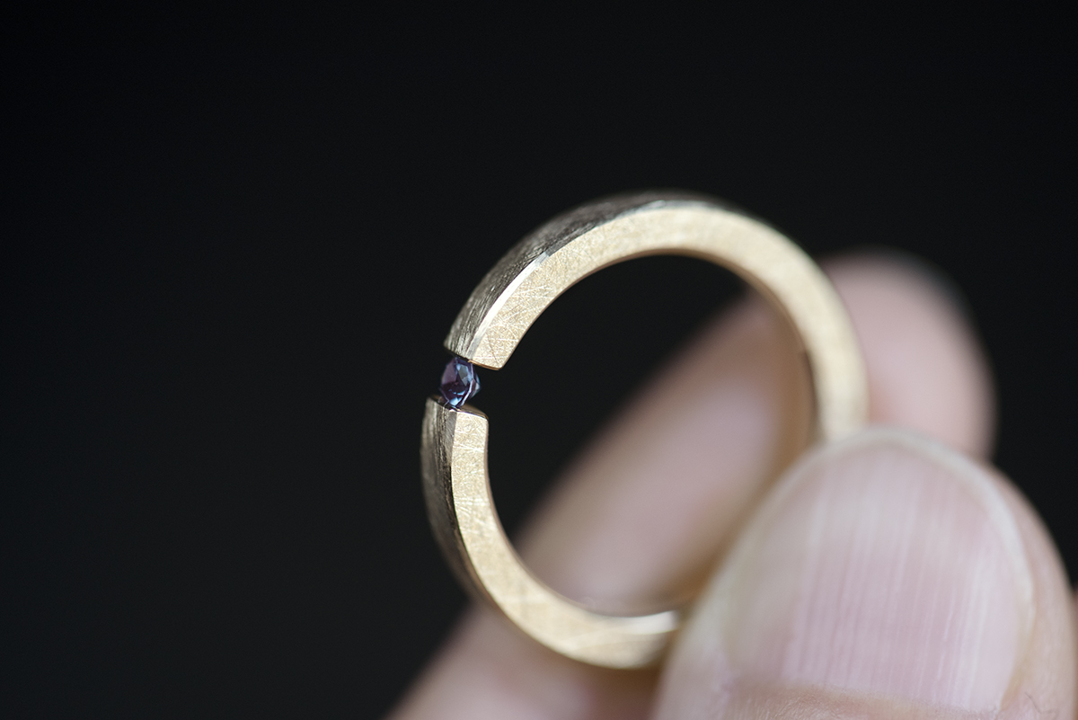 Minimalistyczny pierścionek typu tension ring z żółtego złota próby 585 z syntetycznym zmieniającym kolor aleksandrytem. Końce prostej szyny o przekroju prostokątnym ściskają kamień z obu stron. Pierścionek przypomina prostą obrączkę. Może pełnić funkcję pierścionka zaręczynowego dla kobiety lub mężczyzny lub obrączki. Pierścionek trzymany w palcach.
