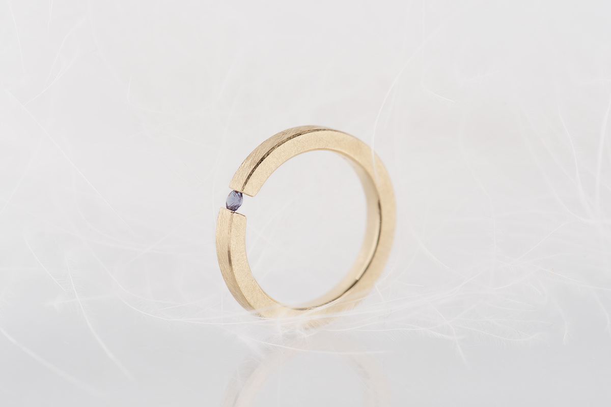 Minimalistyczny pierścionek typu tension ring z żółtego złota próby 585 z syntetycznym zmieniającym kolor aleksandrytem. Końce prostej szyny o przekroju prostokątnym ściskają kamień z obu stron. Pierścionek przypomina prostą obrączkę. Może pełnić funkcję pierścionka zaręczynowego dla kobiety lub mężczyzny lub obrączki.
