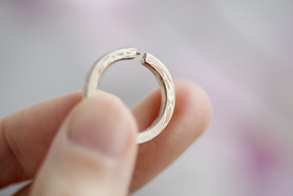 Pierścionek mokume gane srebro i miedź z moissanitem trzymany w palcach. Szyna szerokości i wysokości pierścionka. Srebro i miedź układają się we wzory przypominające słoje drewna.