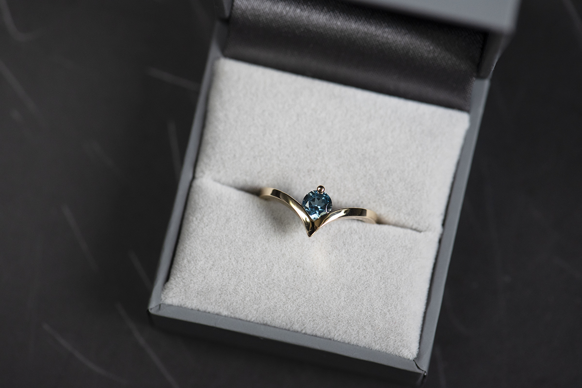 Pierścionek z żółtego złota próby 750 z topazem London blue umieszczony w szarym pudełku na biżuterię. Szyna pierścionka jest wygięta w kształcie litery v. W miejscu wcięcia jest oprawiony niebieski kamień.