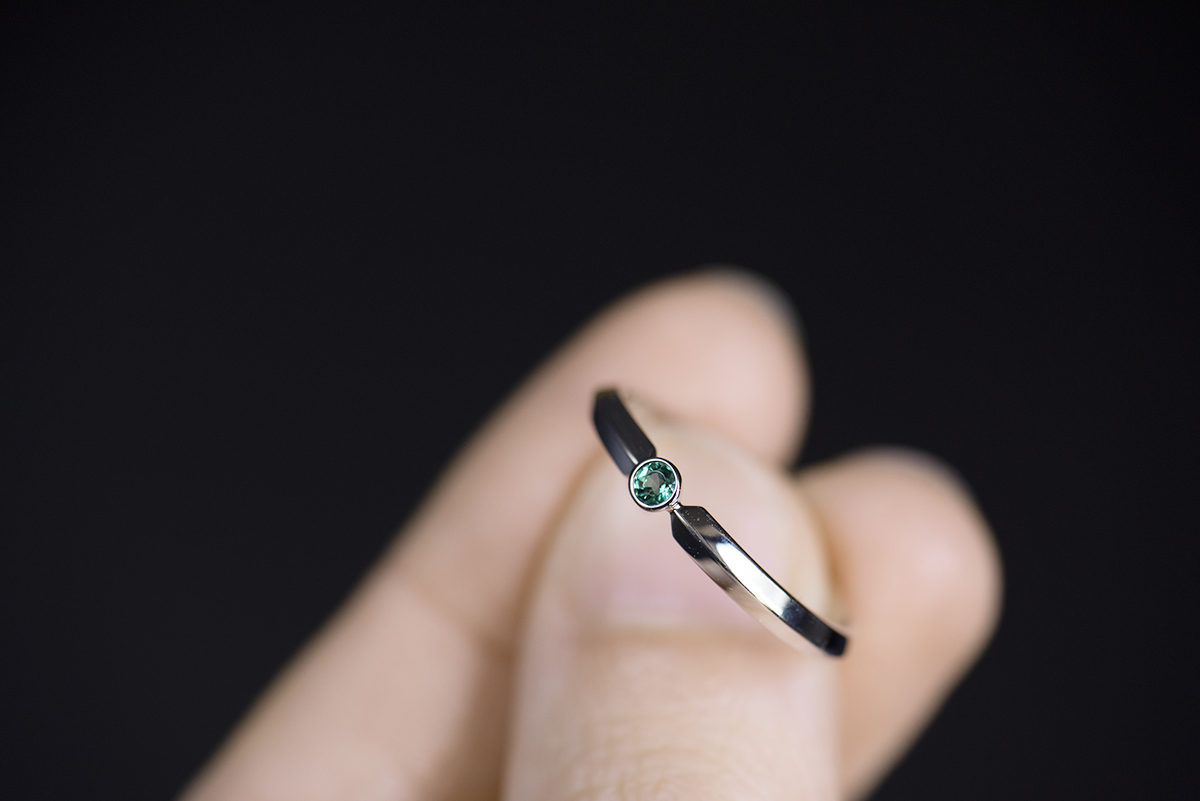 Pierścionek z białego złota próby 585 z syntetycznym szmaragdem. Szyna o profilu trójkątnym zwęża się przy kamieniu tworząc kształt przypominający ostrze miecza. Obrączka jest tej samej szerokości co kamień czyli 2,5 mm. Pierścionek jest trzymany w palcach na czarnym tle.