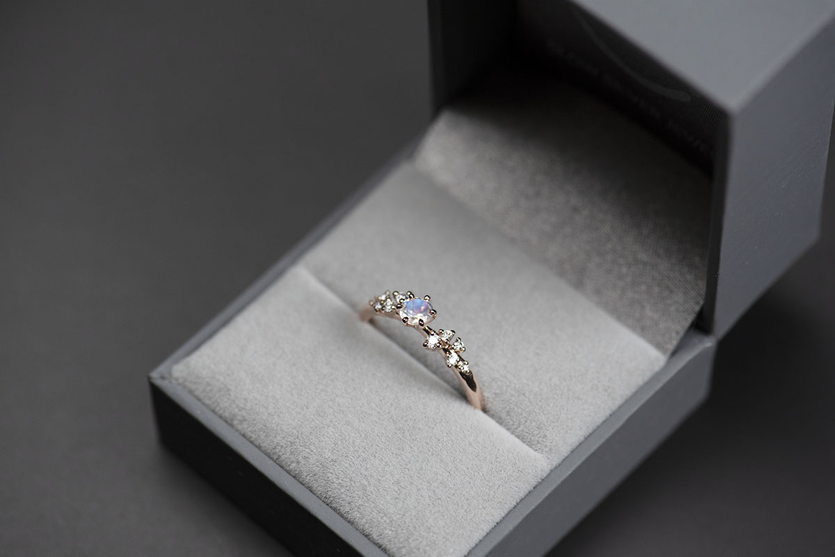 Pierścionek z różowego złota próby 585 z niebieskim kamieniem księżycowym i okrągłymi diamentami po bokach w szarym pudełku na biżuterię. Zdjęcie wykonane w rozproszonym świetle dziennym.