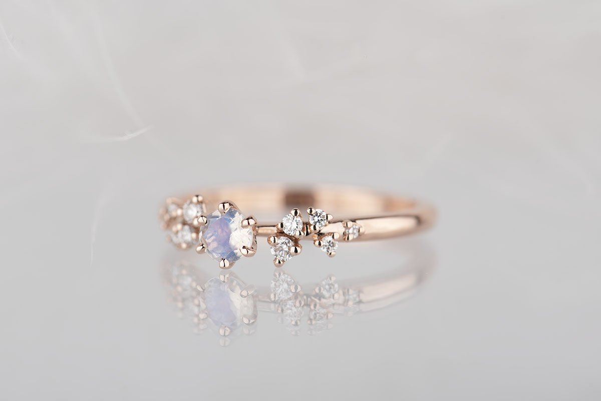 Pierścionek zaręczynowy z różowego złota próby 585 z niebieskim kamieniem księżycowym na środku oraz małymi diamentami po bokach. Fotografia wykonana na białym tle w studio w oświetleniu lamp błyskowych.