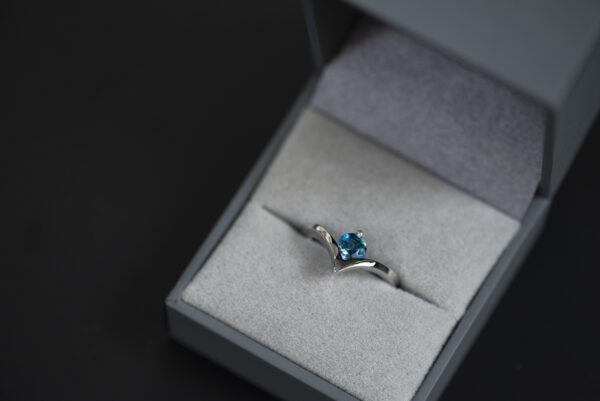 Pierścionek ze srebra 925 z topazem London blue w szarym pudełku na biżuterię. Szyna pierścionka jest wygięta w kształcie litery v. W miejscu wcięcia jest oprawiony niebieski kamień.