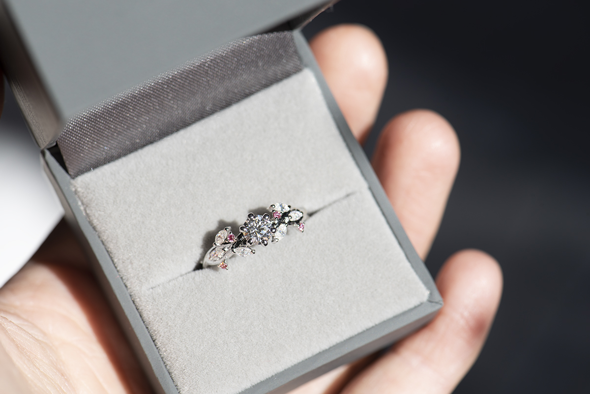 Pierścionek z białego złota próby 585 z diamentem laboratoryjnym 0,4 ct, sybtetycznymi moissanitami markizami i okrągłymi różowymi diamentami. Pierścionek zaręczynowy umieszczony w szarym pudełku na biżuterię.