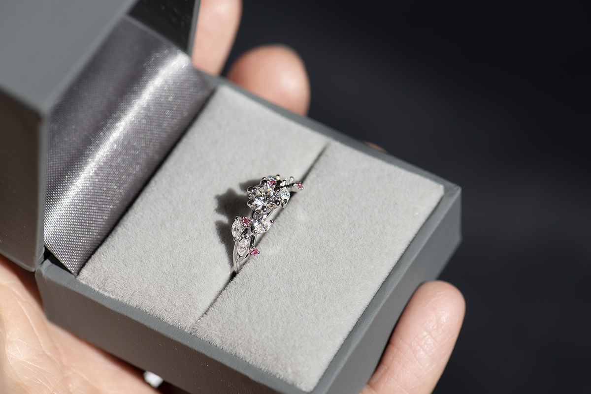 Pierścionek z białego złota próby 585 z diamentem laboratoryjnym 0,4 ct, sybtetycznymi moissanitami markizami i okrągłymi różowymi diamentami. Pierścionek zaręczynowy umieszczony w szarym pudełku na biżuterię.