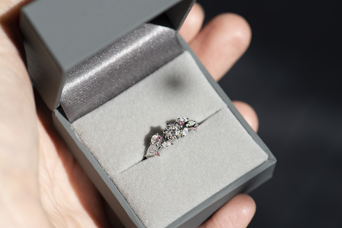 Pierścionek z białego złota próby 585 z diamentem laboratoryjnym 0,4 ct, sybtetycznymi moissanitami markizami i okrągłymi różowymi diamentami. Pierścionek zaręczynowy umieszczony w szarym pudełku na biżuterię.