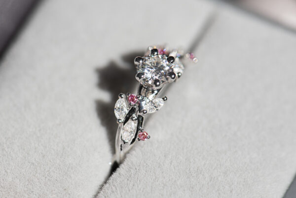 Pierścionek z białego złota próby 585 z diamentem laboratoryjnym 0,4 ct, sybtetycznymi moissanitami markizami i okrągłymi różowymi diamentami. Pierścionek zaręczynowy umieszczony w szarym pudełku na biżuterię, pokazany od boku.
