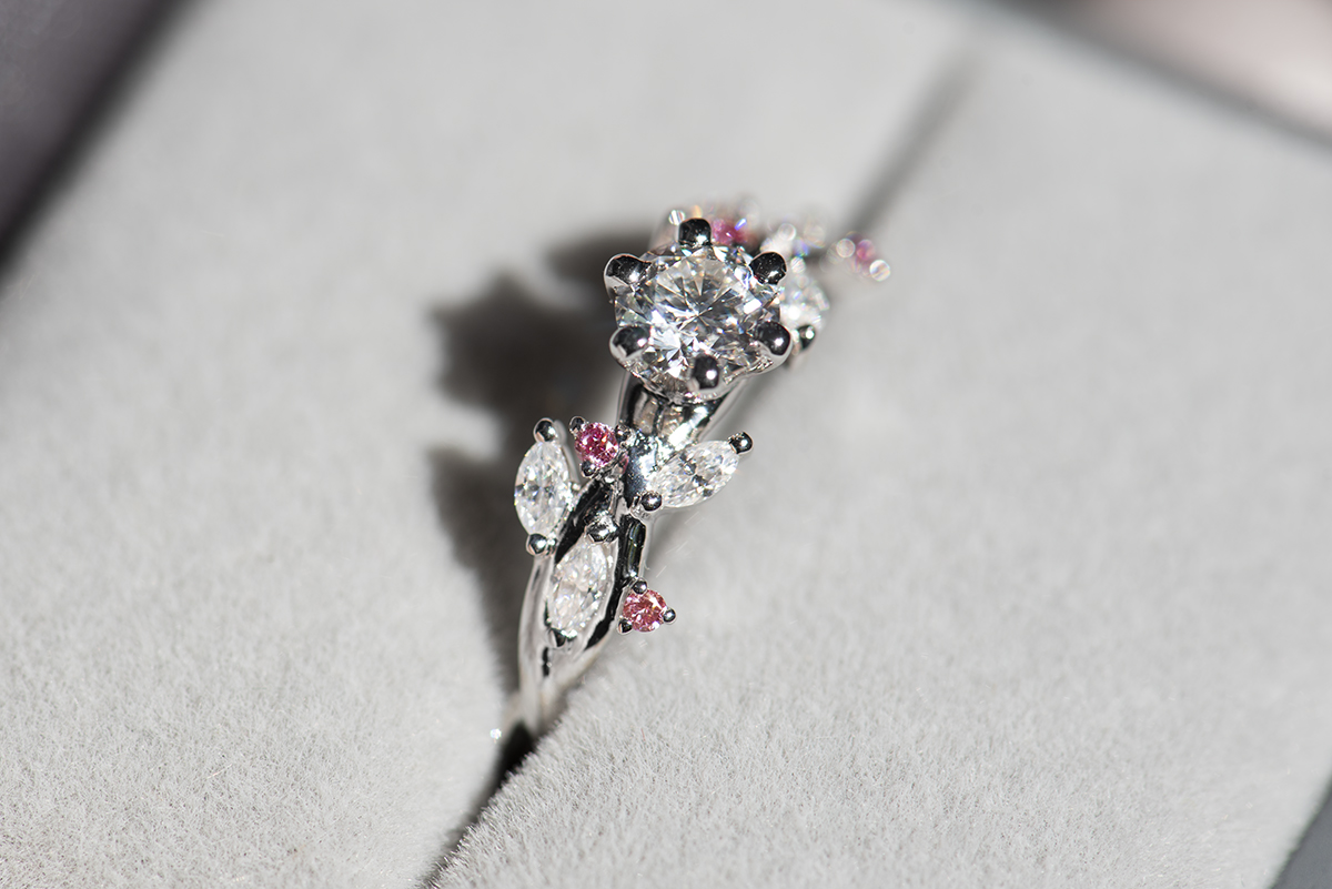 Pierścionek z białego złota próby 585 z diamentem laboratoryjnym 0,4 ct, sybtetycznymi moissanitami markizami i okrągłymi różowymi diamentami. Pierścionek zaręczynowy umieszczony w szarym pudełku na biżuterię, pokazany od boku.