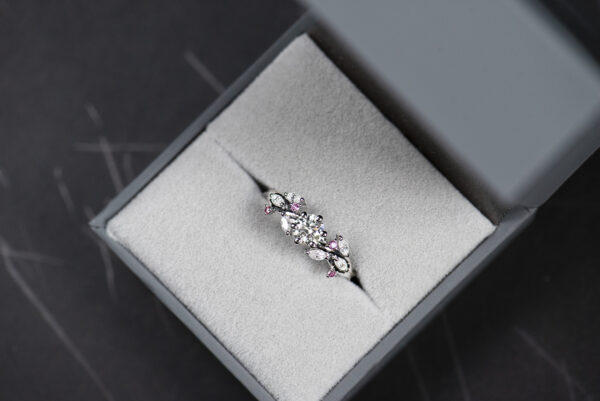 Pierścionek z białego złota próby 585 z diamentem laboratoryjnym 0,4 ct, sybtetycznymi moissanitami markizami i okrągłymi różowymi diamentami. Pierścionek zaręczynowy umieszczony w szarym pudełku na biżuterię.