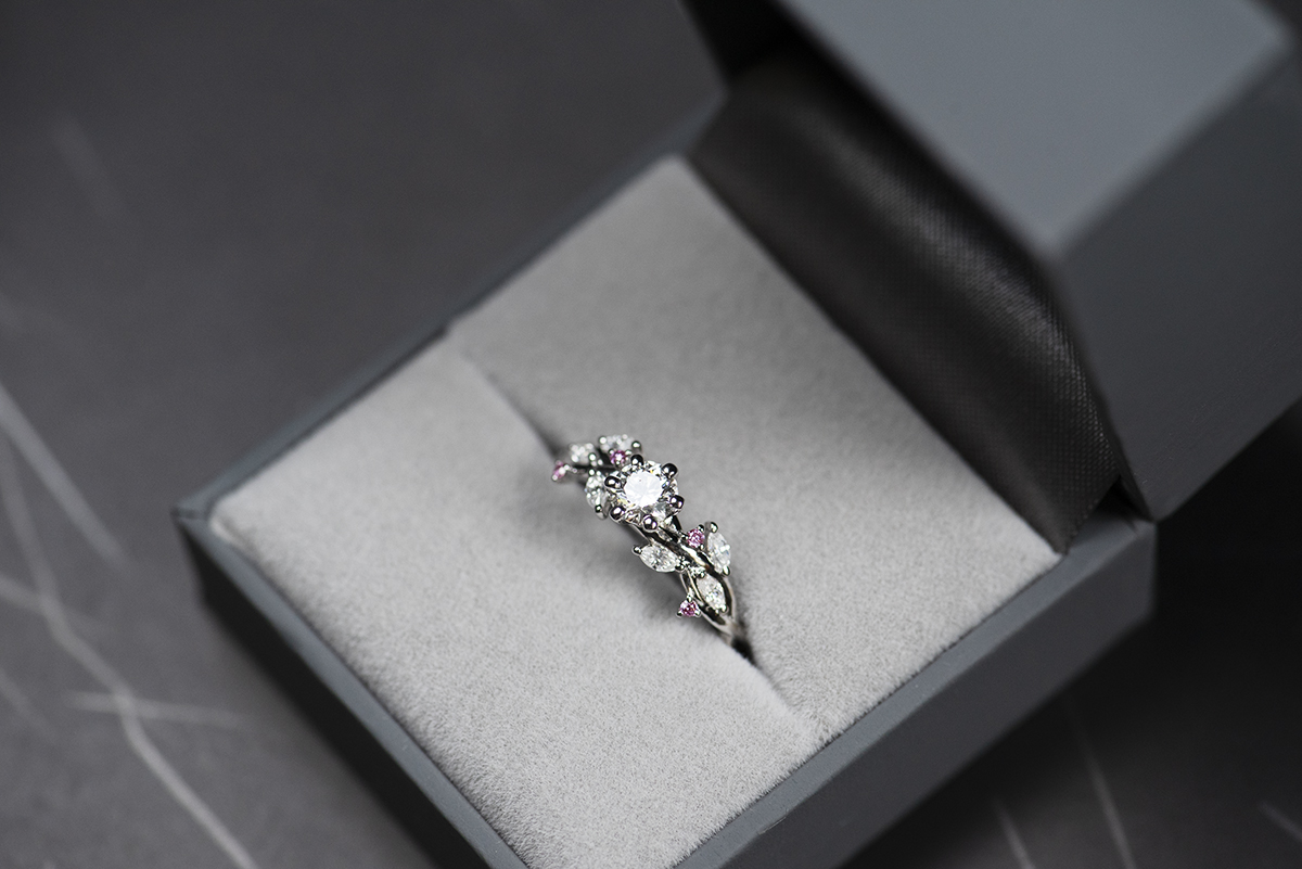 Pierścionek z białego złota próby 585 z diamentem laboratoryjnym 0,4 ct, sybtetycznymi moissanitami markizami i okrągłymi różowymi diamentami. Pierścionek zaręczynowy umieszczony w szarym pudełku na biżuterię.