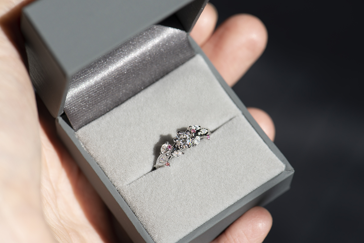 Pierścionek z białego złota próby 585 z diamentem laboratoryjnym 0,4 ct, sybtetycznymi moissanitami markizami i okrągłymi różowymi diamentami. Pierścionek zaręczynowy umieszczony w szarym pudełku na biżuterię.