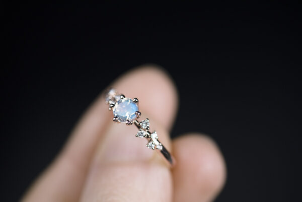 Pierścionek z żółtego złota próby 585 z niebieskim kamieniem księżycowym i małymi diamentami po bokach trzymany w palcach. Zdjęcie wykonane w rozproszonym świetle dziennym