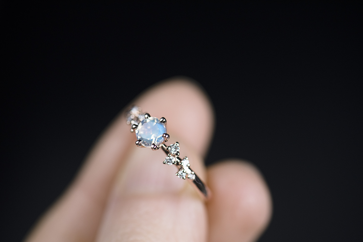 Pierścionek z żółtego złota próby 585 z niebieskim kamieniem księżycowym i małymi diamentami po bokach trzymany w palcach. Zdjęcie wykonane w rozproszonym świetle dziennym