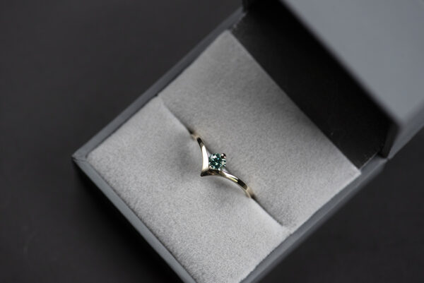 Pierścionek ze złota próby 585 z syntetycznym moissanitem szmaragdowym umieszczony w szarym pudełku na biżuterię. Szyna wygięta w kształcie litery V. W miejscu wygięcie jest oprawiony kamień o średnicy 4 mm (0,3 ct).