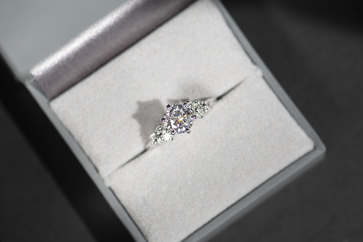 Pierścionek zaręczynowy z białego złota z diamentem 1 ct oraz drobnymi diamentami po bokach. Pierścionek umieszczony w szarym pudełku na biżuterię. Zdjęcie w naturalnym oświetleniu