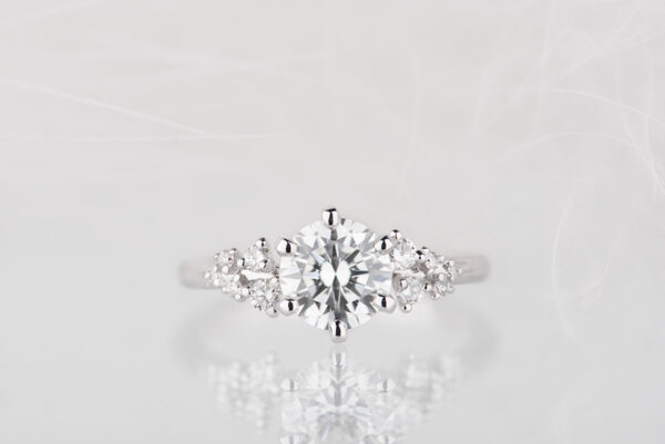 Pierścionek zaręczynowy z białego złota z diamentem 1 ct oraz drobnymi diamentami po bokach. Zdjęcie wykonane na jasnym tle.