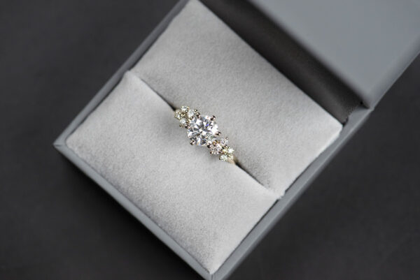 Pierścionek zaręczynowy z żółtego złota z diamentem 1 ct oraz drobnymi diamentami po bokach. Pierścionek umieszczony w szarym pudełku na biżuterię. Zdjęcie w naturalnym oświetleniu