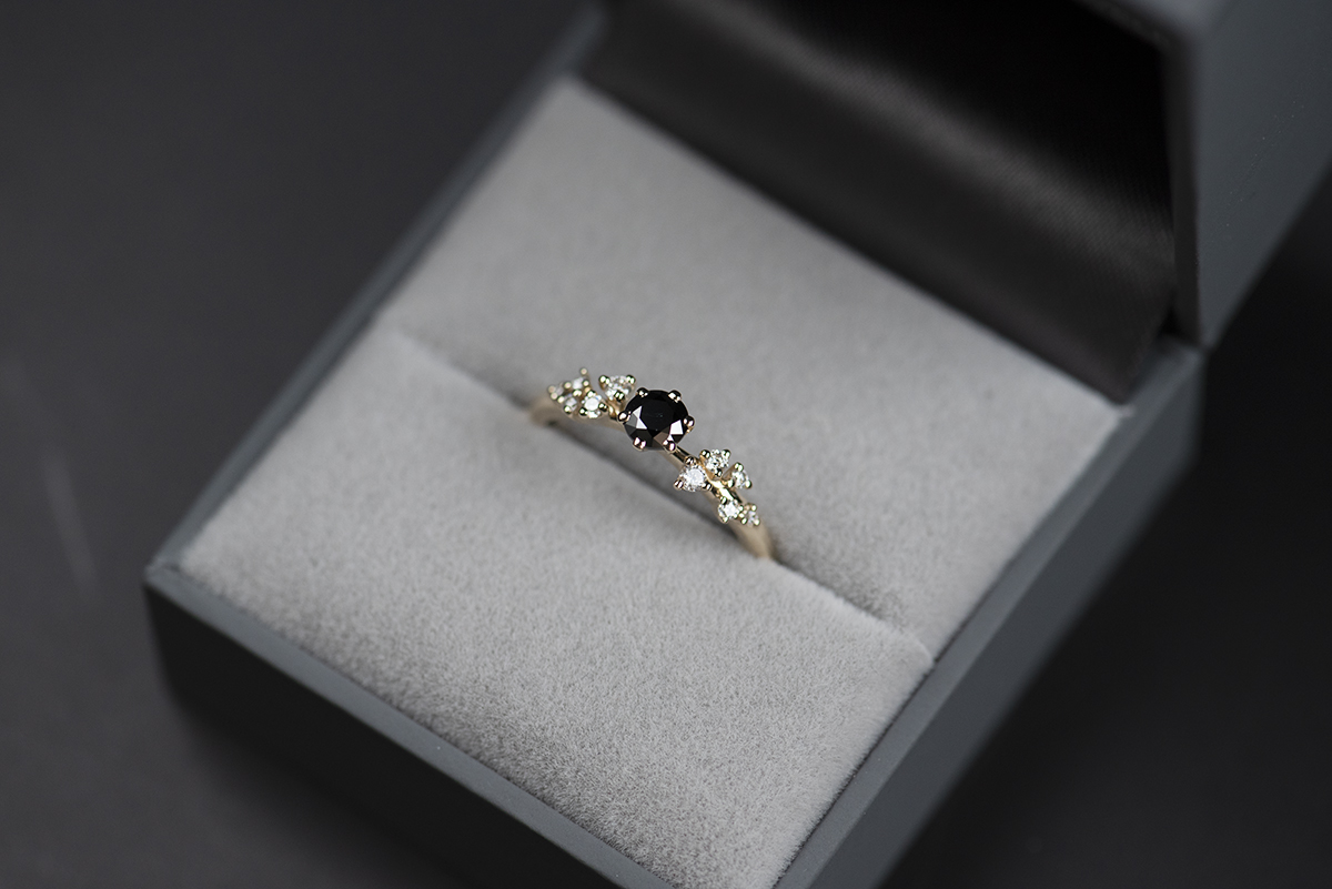 Pierścionek zaręczynowy ze złota próby 585 z czarnym diamentem i drobnymi białymi diamentami po bokach umieszczony w szarym pudełku na biżuterię. Zdjęcie wykonane w naturalnym oświetleniu