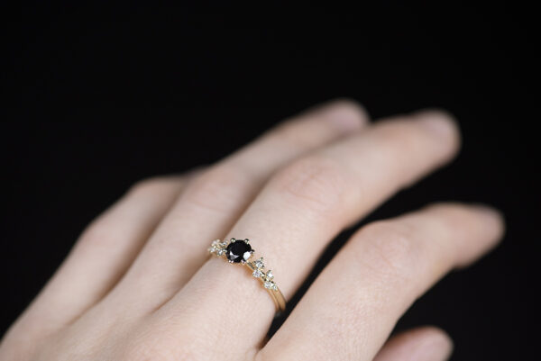 Pierścionek zaręczynowy ze złota próby 585 z czarnym diamentem 0,5 karata i drobnymi diamentami po bokach. Pierścionek sfotografowany na palcu. Zdjęcie wykonane w świetle dziennym
