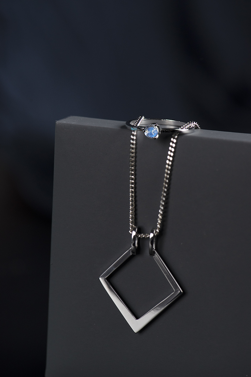 Ring holder wykonany z białego złota próby 585. Zawieszka na którą można założyć pierścionek bez konieczności rozpinania łańcuszka.Łańcuszek przewieszony przez szare pudełko na biżuterię. Na pokrywie pudełka umieszczony pierścionek z białego złota z kamieniem księżycowym.