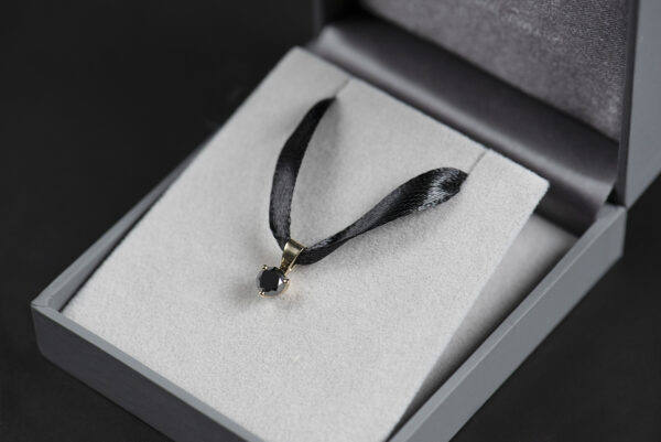 Zawieszka ze złota 750 z czarnym diamentem 0,5 ct zawieszona na czarnej wstążeczce w szarym pudełku na biżuterię.