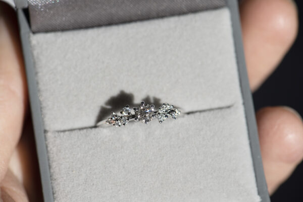 pierścionek zaręczynowy z białego złota z głównym kamieniem diamentem sól i pieprz o macie 0,3 karata i oprawionymi po bokach białymi diamentami. Pierścionek umieszczony w szarym pudełku na biżuterię. Zdjęcie wykonane w bezpośrednim świetle słonecznym