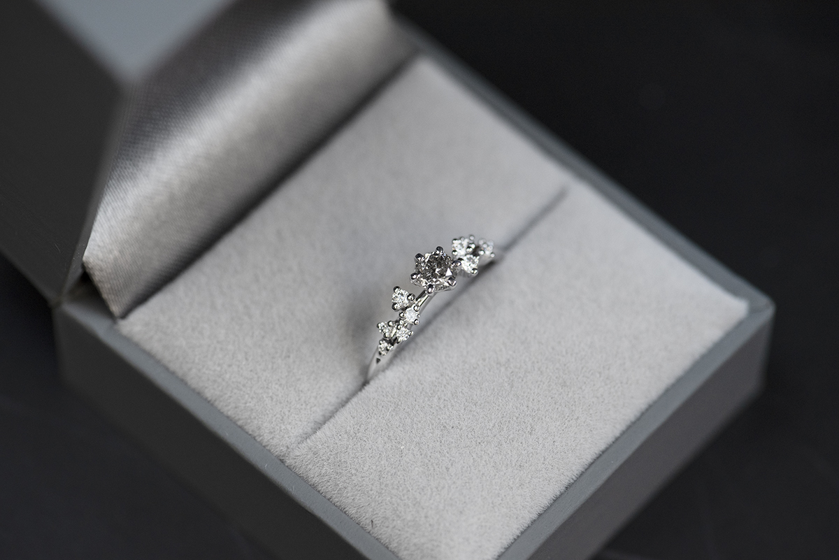 pierścionek zaręczynowy z białego złota z głównym kamieniem diamentem sól i pieprz o macie 0,3 karata i oprawionymi po bokach białymi diamentami. Pierścionek umieszczony w szarym pudełku na biżuterię. Zdjęcie wykonane w rozproszonym świetle