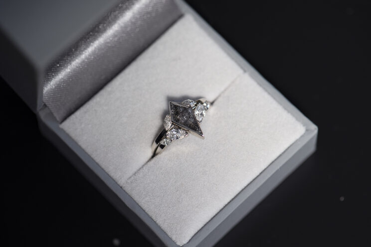 Pierścionek z białego złota z diamentem sól i pieprz w szlifie kite. Po bokach głównego kamienia po jednym diamencie sol i pieprz w szlifie markiza. Pierścionek umieszczony w szarym pudełku na biżuterię razem z pasującą do niego obrączką również z markizami diamentami sól i pieprz.